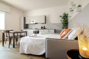 Una habitación de hotel moderna con cama, sofá, mesa de comedor con sillas, kitchenette y un gran ventanal.