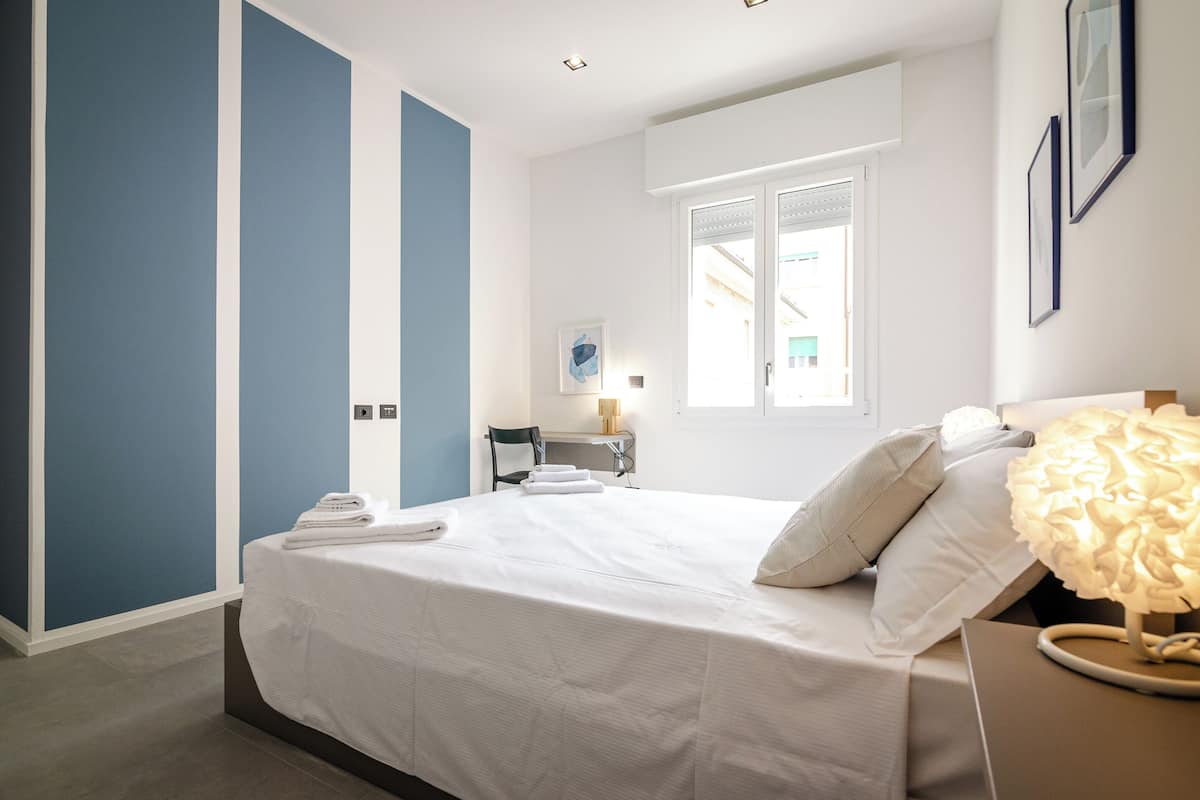 A hotel room with a bed, a desk, a chair, a window, and a lamp.