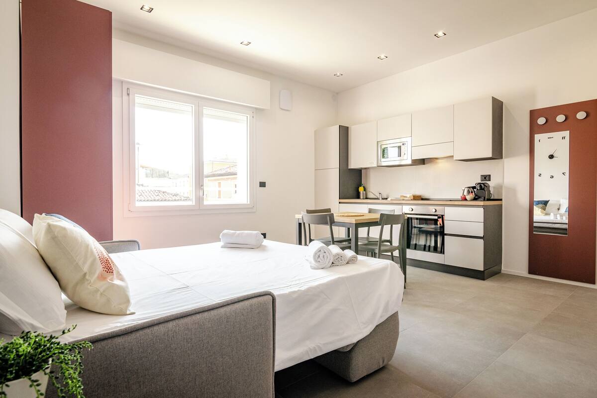 A modern hotel room with a bed, a small kitchenette, a dining area, and a view of the city.