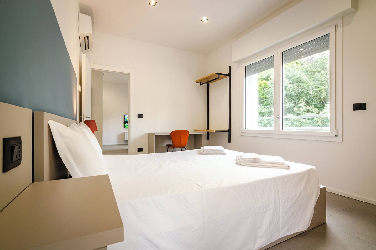 A modern hotel room with a bed, a desk, an orange chair, and a large window.