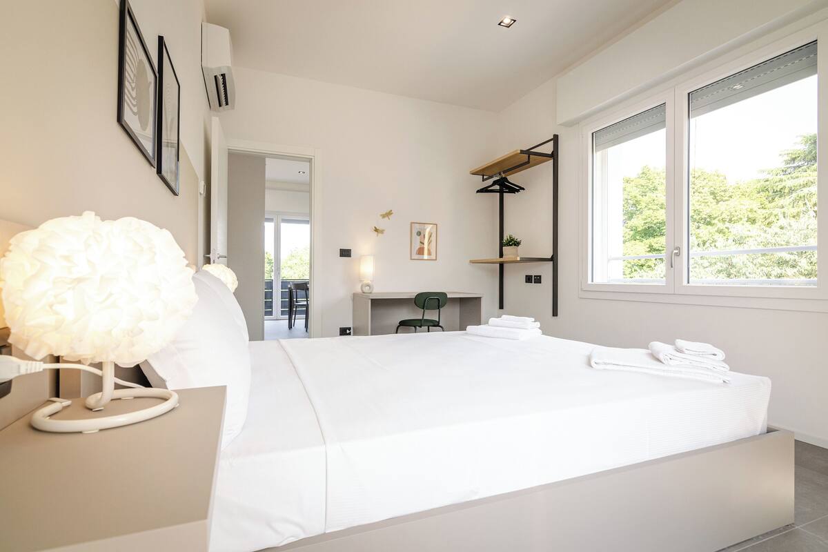 A modern bedroom with a large bed, a desk with a lamp, a chair, a shelf, and a window with a view of greenery.
