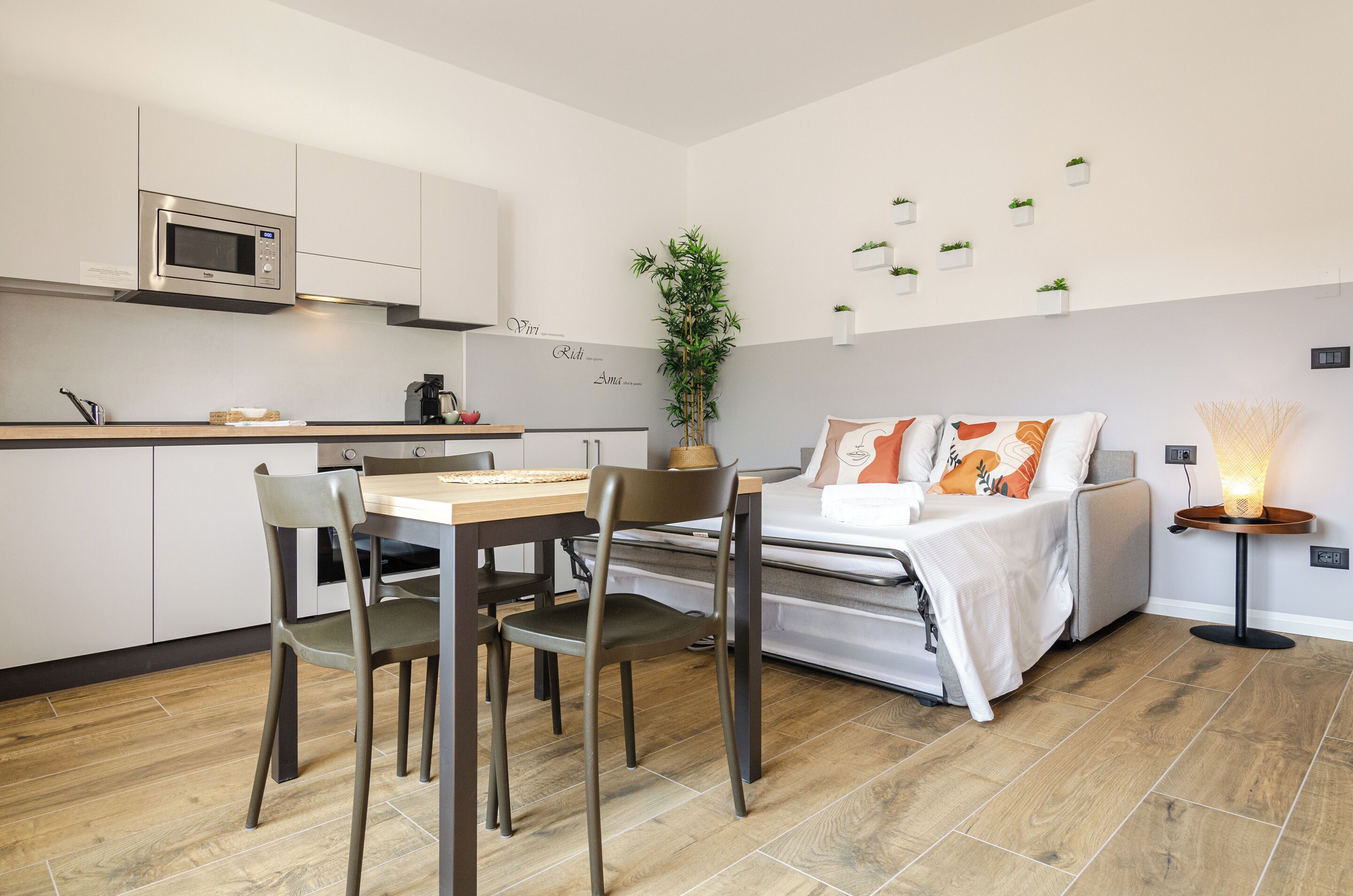 Een knus woongedeelte met kitchenette, een eethoek, een bed met nachtkastje, en een kleine tafel met lamp.