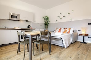 Un espacio habitable compacto con kitchenette, zona de comedor, cama con mesita de noche y una pequeña mesa con lámpara.