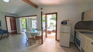 Een ruime woonkamer met een eettafel, stoelen, een keuken met een koelkast en magnetron, en een schuifpui naar een terras.