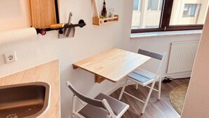 Kompakti keittiö, jossa on puinen pöytä ja tuolit, pieni hylly pulloille ja ikkuna, josta avautuu näkymä rakennuksiin.