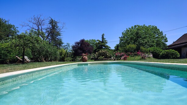 A clear swimming pool with a tiled edge, surrounded by lush greenery and a house in the background. - Holiday Home "Maison 6 à 10 personnes" with Private Pool, Terrace and Wi-Fi (Sainte-Croix)