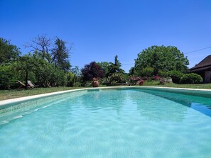 A clear swimming pool with a tiled edge, surrounded by lush greenery and a house in the background.