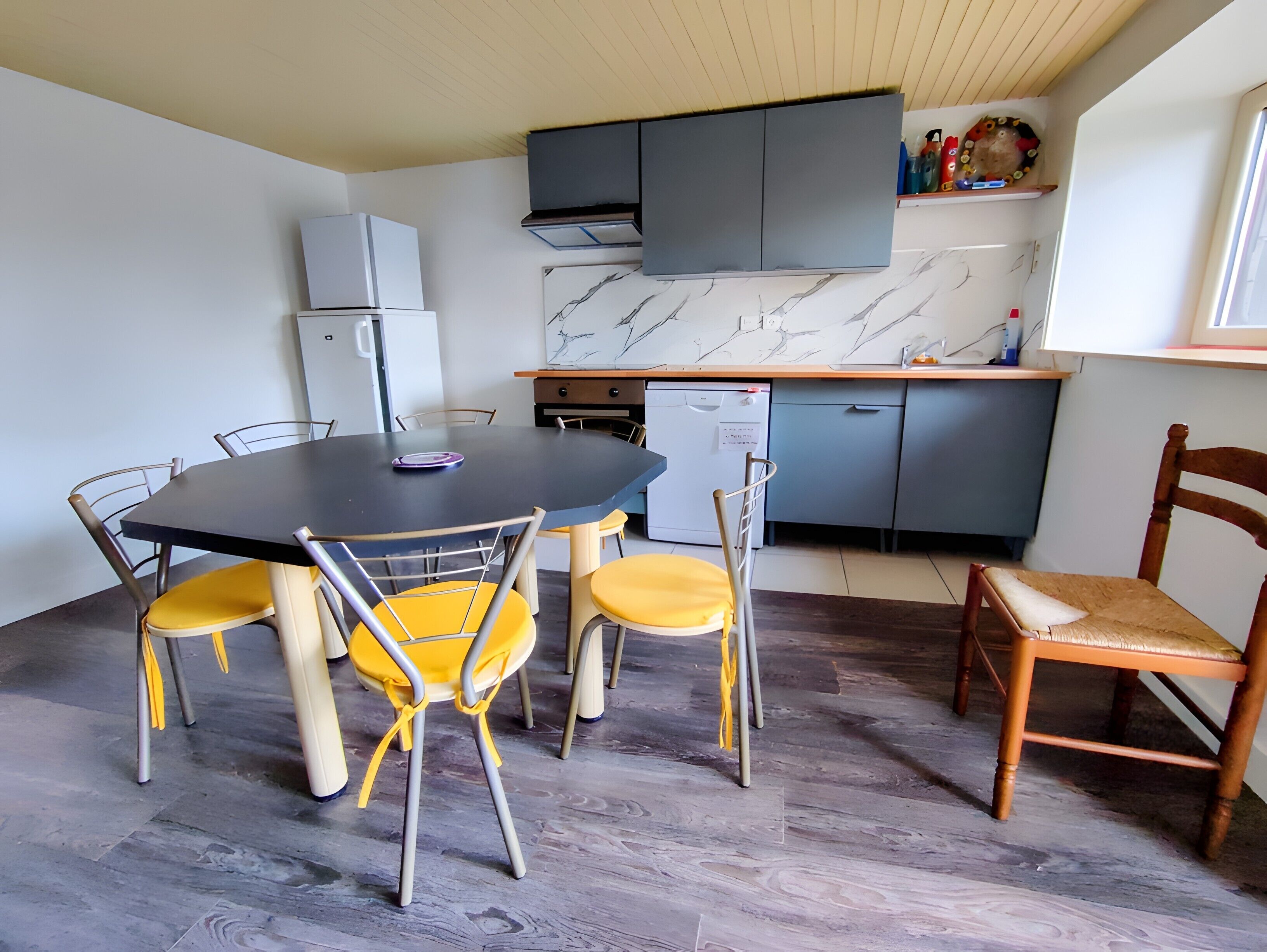 A compact kitchen with a built-in oven, a small refrigerator, and a microwave. There is a dining table with chairs and a wooden chair with a cushion.