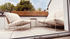 A balcony with two white cushioned chairs and a small table, a wooden planter, and a view of trees and buildings.