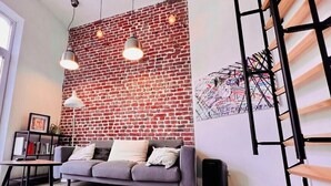 A living room with a brick wall, a grey sofa, a black speaker, and a ladder against the wall.