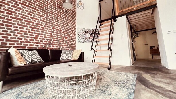 A room with a brick wall, a black leather sofa, a round white table, and a ladder leading to an upper level.