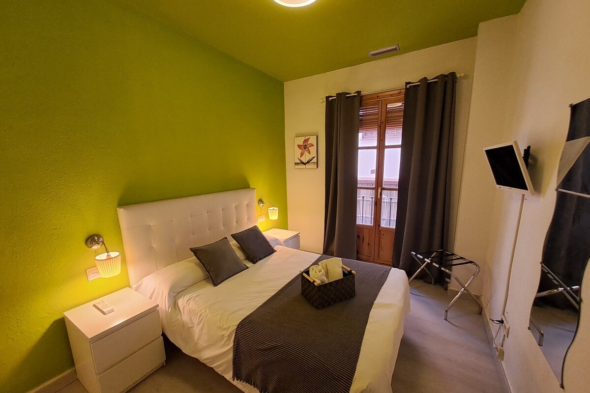 Ein Hotelzimmer mit grüner Wand, einem weißen Bett, einem Nachttisch mit Lampe, einem Fenster mit Vorhängen und einem an der Wand montierten Fernseher.