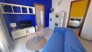 Un salon avec un canapé, un téléviseur, des étagères blanches et un lit à l’arrière-plan.
