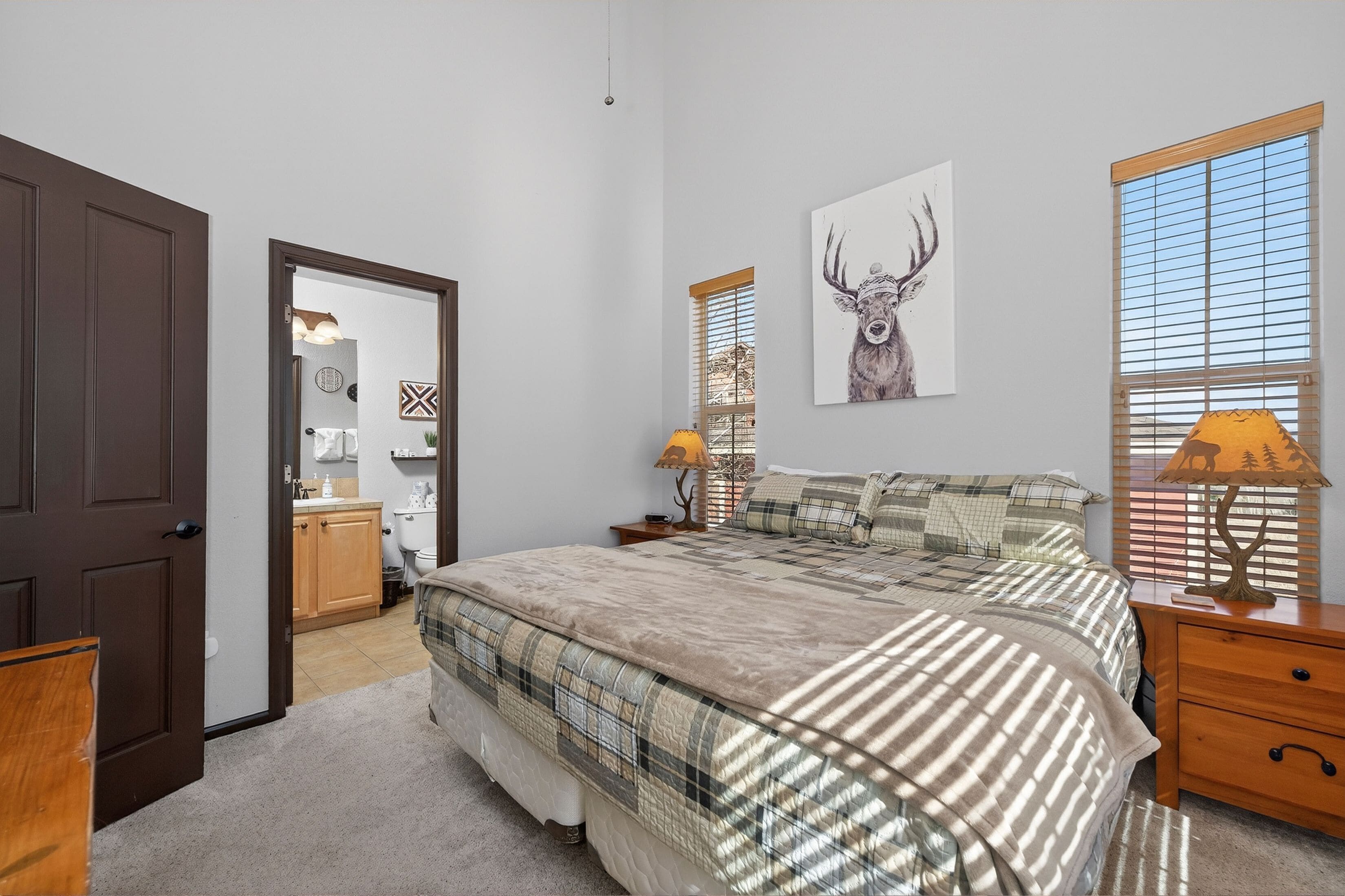 A bedroom with a large bed, a wooden nightstand, a lamp, a painting of a deer, and a window with blinds.