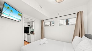 Et hotelværelse med en seng, et fjernsyn ophængt på væggen, et vindue med gardiner og udsigt til stranden.