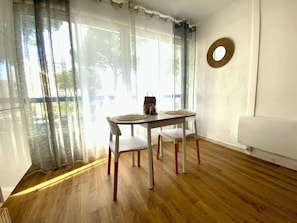 Une pièce avec un plancher en bois, une table ronde entourée de chaises, un canapé blanc et des rideaux légers.