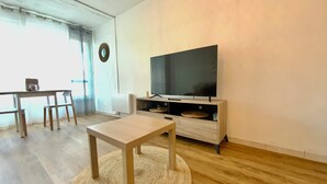 Olohuoneessa on taulutelevisio, puinen sohvapöytä, ruokapöytä tuoleineen ja suuri ikkuna, jossa on verhot.