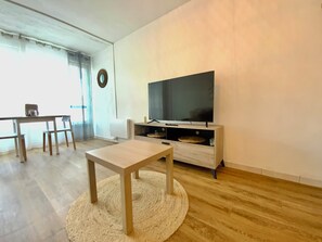 Un salon équipé d’un téléviseur à écran plat, d’une table basse en bois, d’une table à manger avec des chaises et d’une grande fenêtre avec des rideaux.