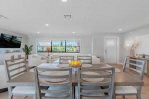 A dining area with a wooden table and chairs, a bowl of fruit, and a view of the outdoors through large windows. - 🏖️Private Heated Pool | Canal Front | Gorgeous Views | Barefoot Escape🏖️ (Fort Myers Beach)