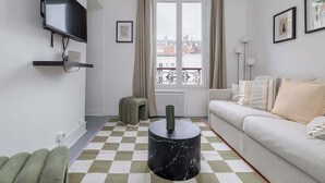 A living room with a sofa, a TV mounted on the wall, a window with curtains, and a checkered floor. - Orleans  - Paris Neuilly 4Pax (Neuilly-sur-Seine)