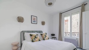 A bedroom with a bed, a bedside table, a chair, a window with curtains, and a wall-mounted fan. - Orleans  - Paris Neuilly 4Pax (Neuilly-sur-Seine)