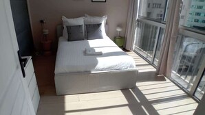 Une chambre à coucher avec un lit, des oreillers, une table de chevet, une lampe et une fenêtre donnant sur des immeubles.