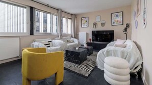 Un salon moderne avec un canapé, un fauteuil jaune, une table basse noire, un téléviseur et des œuvres d’art accrochées aux murs.
