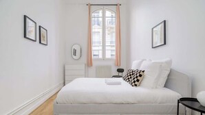 A bedroom with a bed, a radiator, a window with curtains, and framed pictures on the walls. - Perretti - Spacious Flat 2BDR Paris Neuilly (Neuilly-sur-Seine)