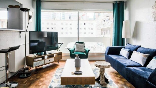Een moderne woonkamer met een flatscreen televisie, een bank, een salontafel en uitzicht op gebouwen door grote ramen.