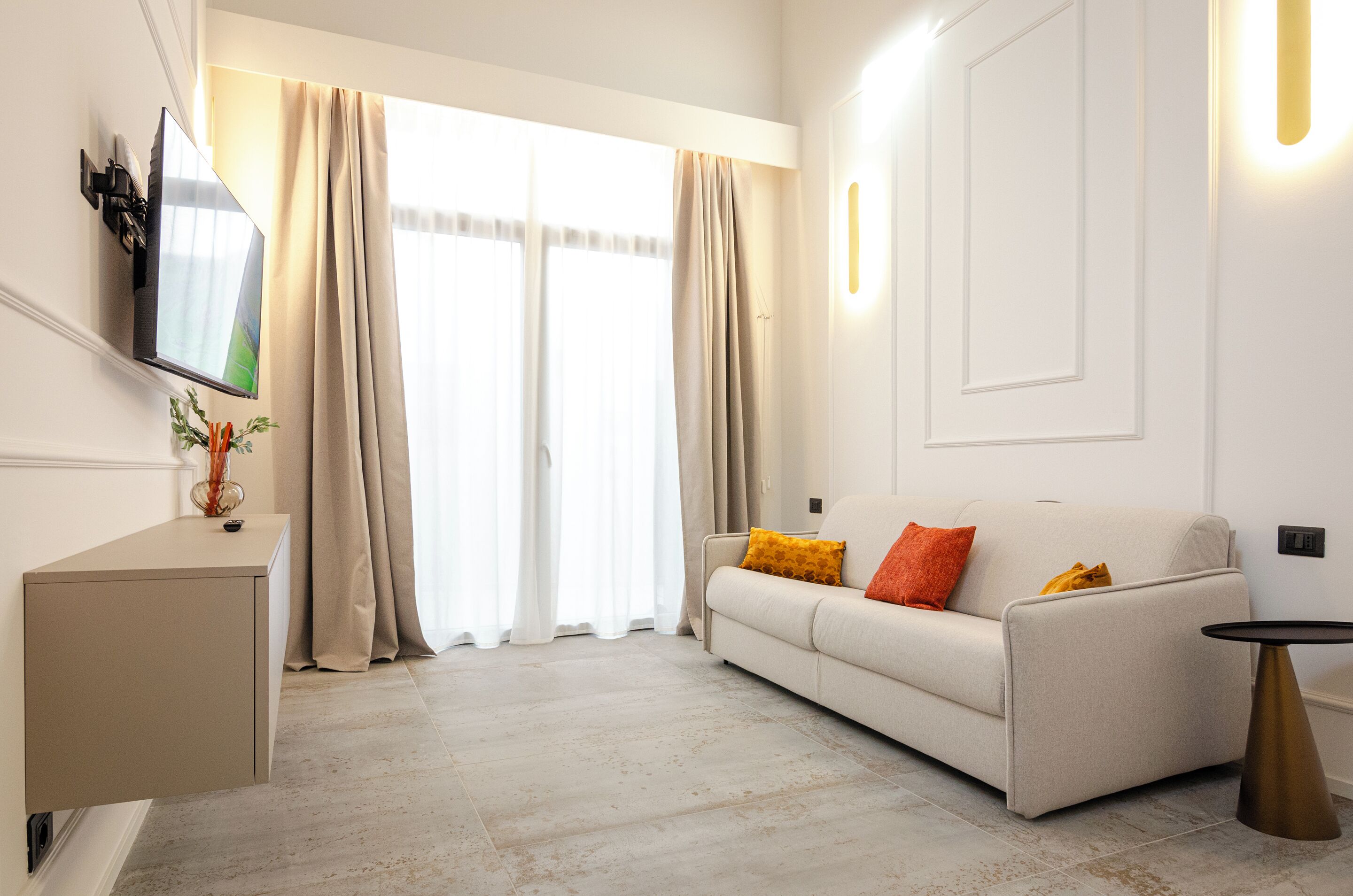 A modern living room with a sofa, a small side table, a mounted TV, and a large window with curtains.