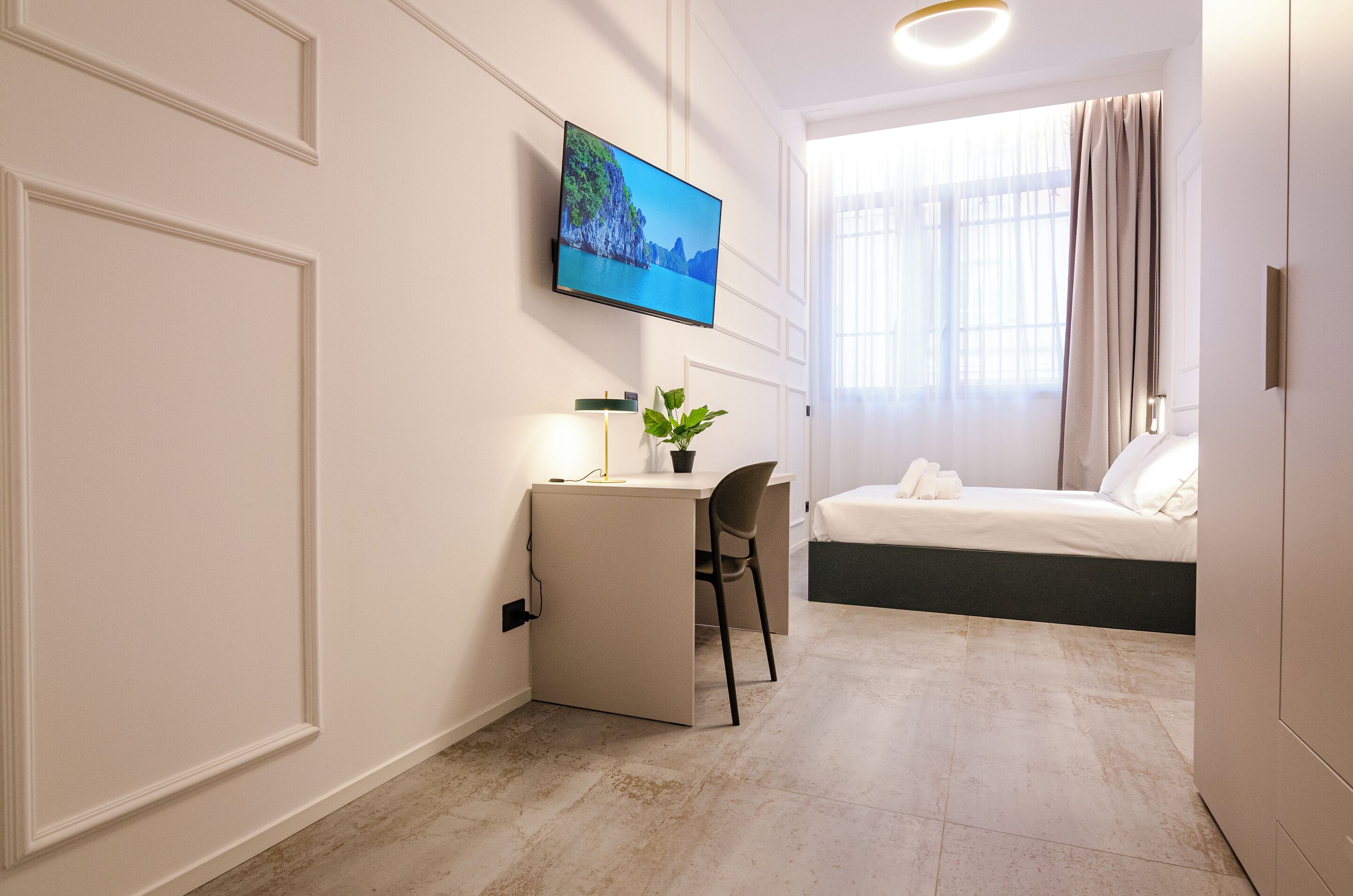Une chambre d’hôtel avec un lit, un bureau et une chaise, un téléviseur fixé au mur et une fenêtre avec des rideaux.
