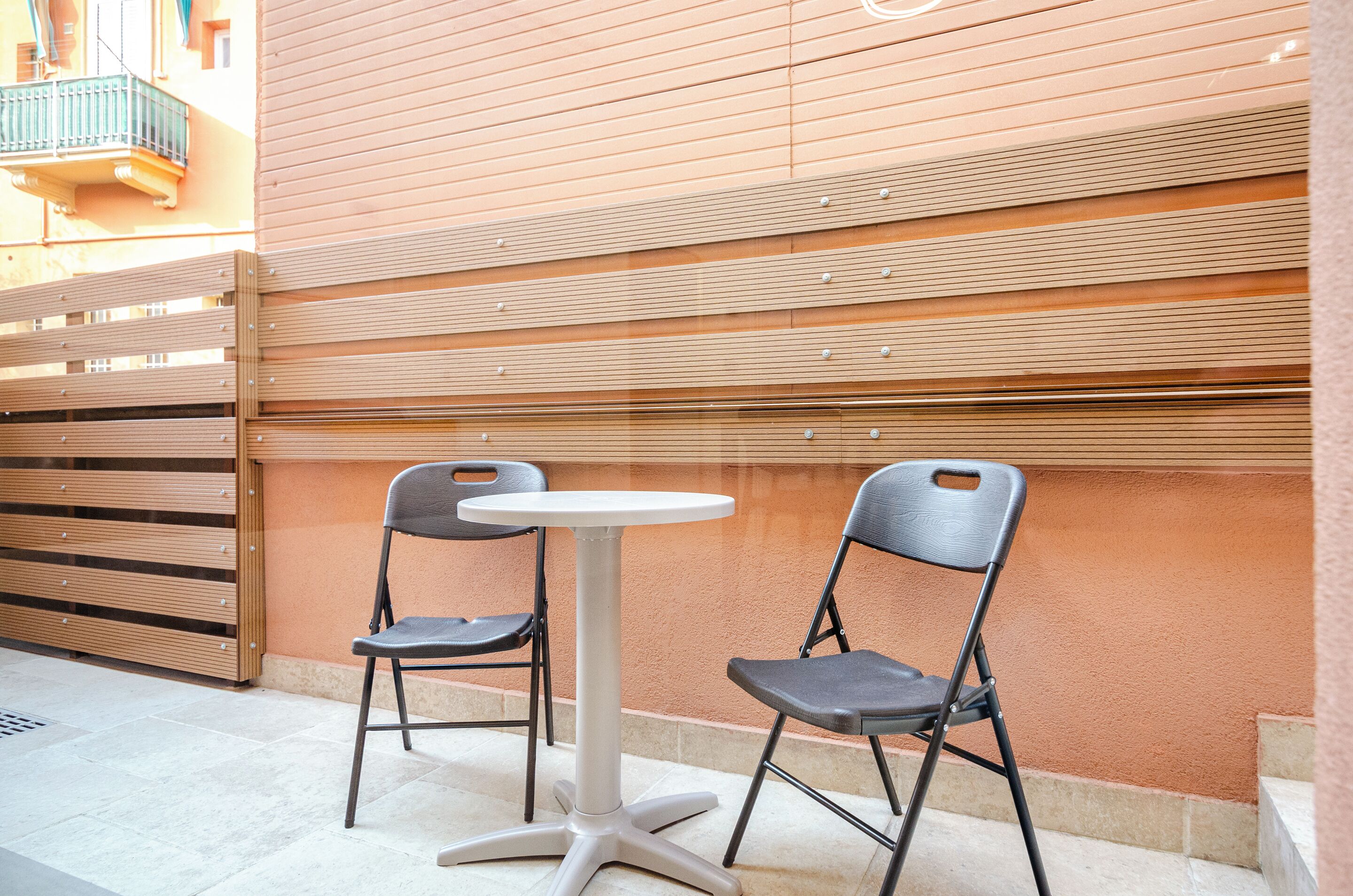 Uma pequena área externa com espaço para sentar, incluindo uma mesa redonda e duas cadeiras, cercada por um muro de madeira, com um edifício de revestimento horizontal.
