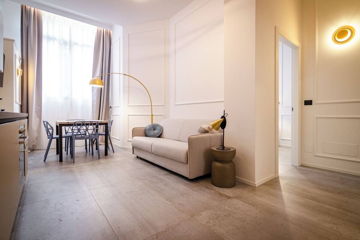 A modern living room with a sofa, a dining table with chairs, a lamp, and a small side table.