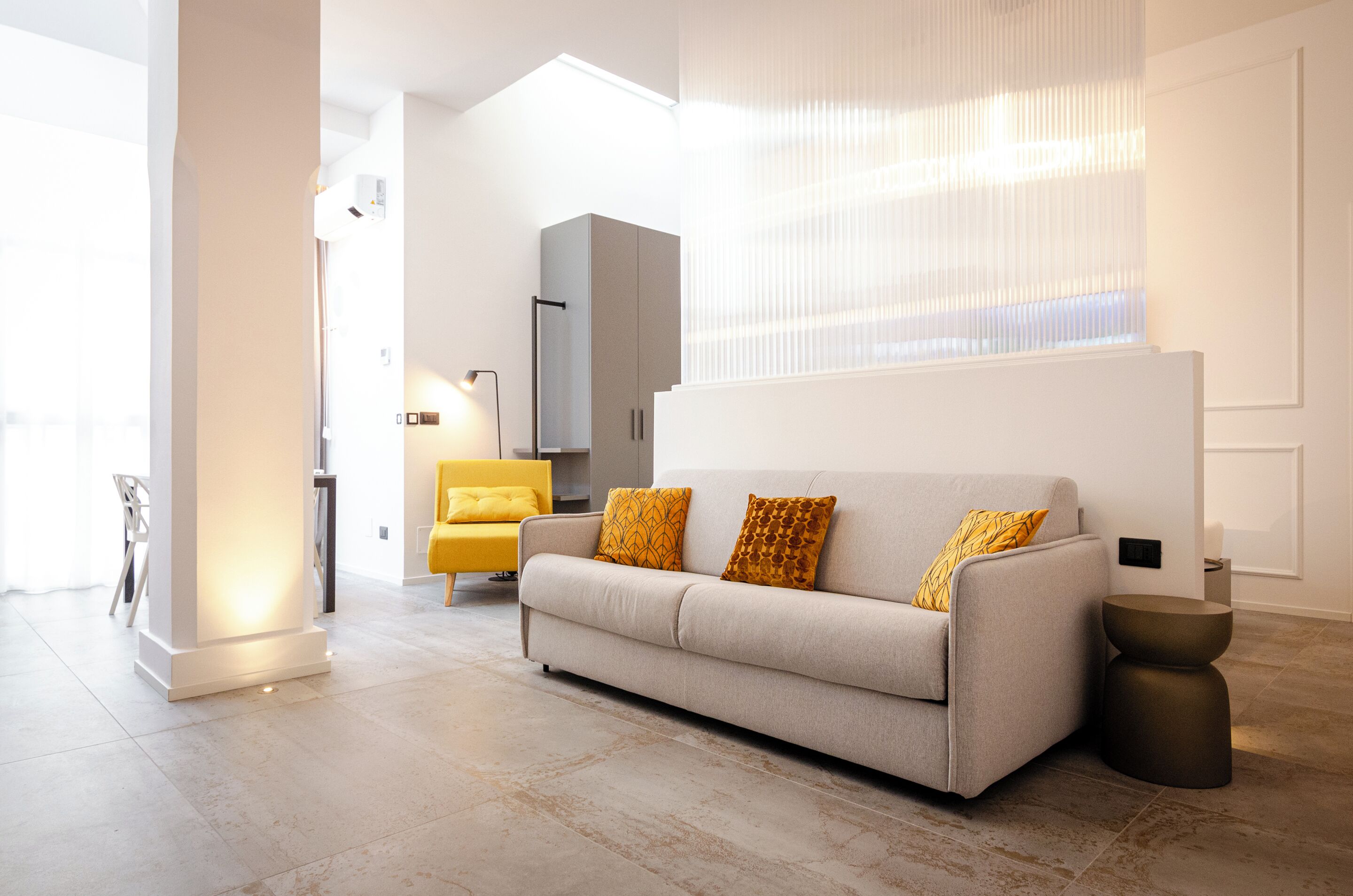 A modern living room with a sofa, a yellow armchair, a tall column, and a large window with blinds.