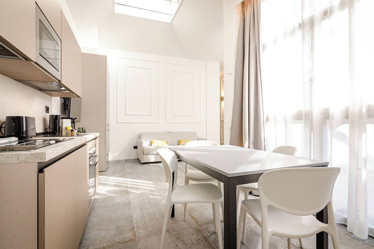A modern kitchen with a dining area, white furniture, and a large window with curtains.