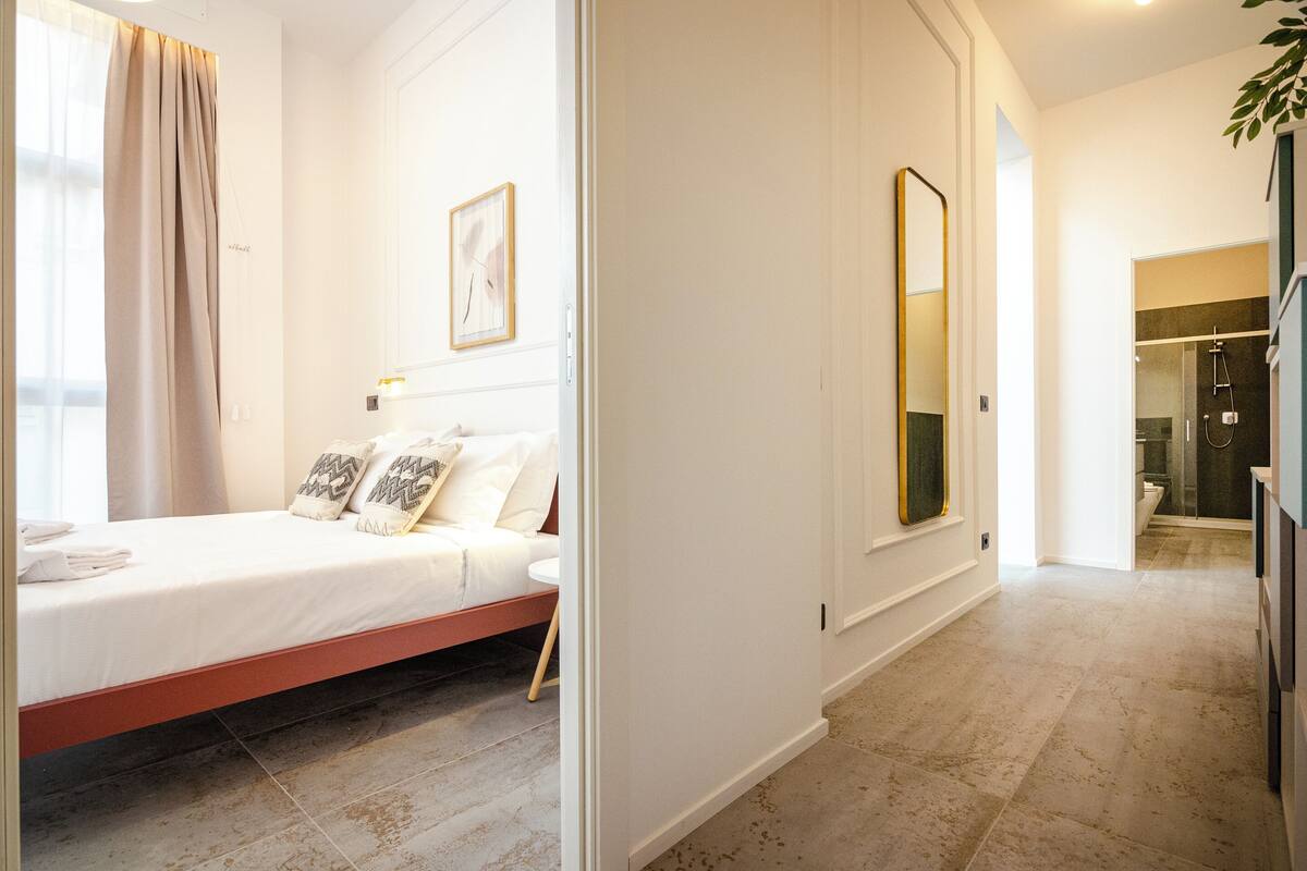 A bedroom with a bed, a bedside table, a mirror, and a bathroom visible through an open door.