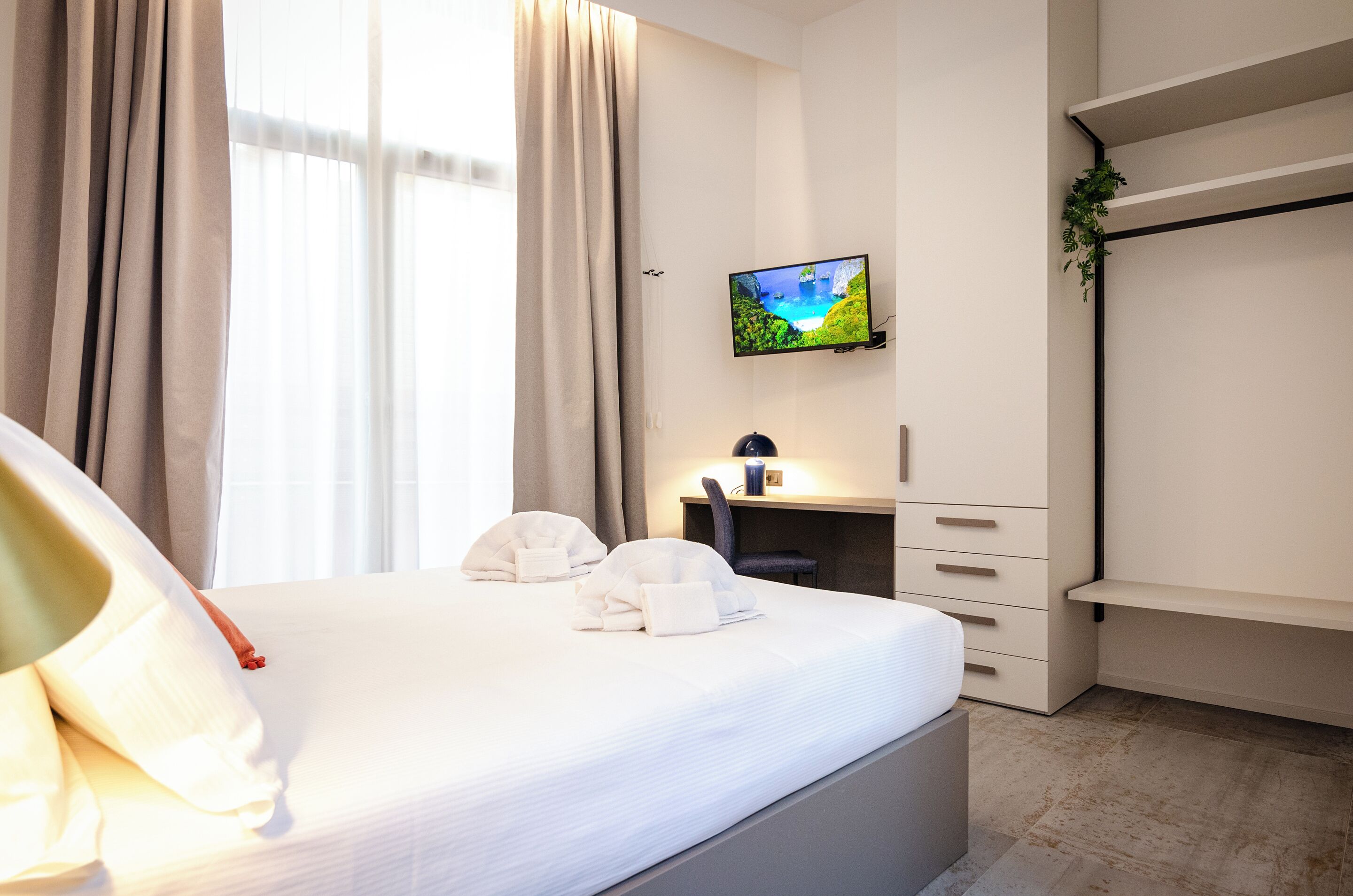 Een hotelkamer met een bed, een aan de muur bevestigde televisie, een bureau met een stoel en een plant.