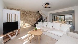 A modern living room with a stone wall, a white sofa, a TV on a wooden stand, and a ceiling fan. - Holiday Home 'Refuge du Sentier Sauvage' with Private Terrace (Belz)