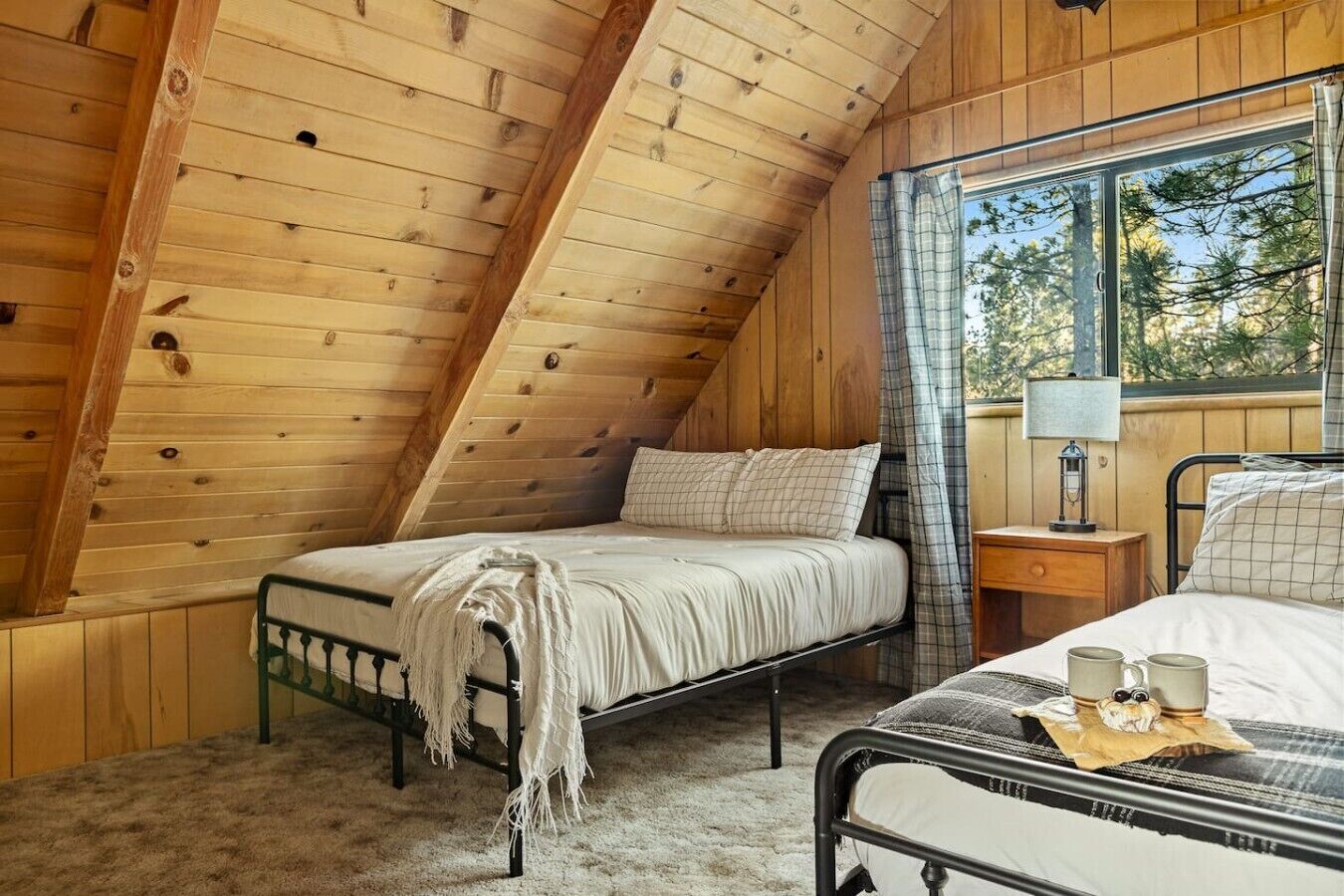 A cozy attic bedroom with a sloped ceiling, a bed with a white comforter, a nightstand with a lamp, and a window with a view of trees.