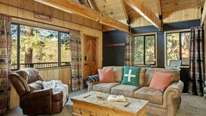 A cozy cabin interior with a leather armchair, a sofa with patterned cushions, a wooden coffee table, and large windows offering a view of trees. - The Silver Coyote, w/ Bear Mtn Views & Firepit (Big Bear Lake)