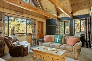 Ein gemütliches Cabin-Interieur mit Ledersessel, einer Couch mit gemusterten Kissen, einem Holztisch und großen Fenstern mit Blick auf Bäume.