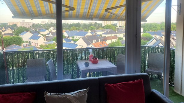 A balcony with a yellow-striped awning, a table with a red pot, and a view of residential buildings. - Large T5 for 7 people (Angers)