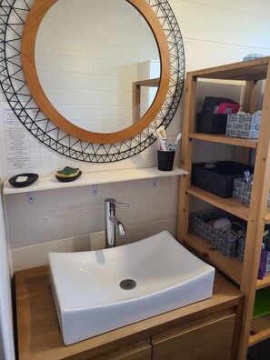 A bathroom with a white sink, a wooden vanity, a mirror with a geometric frame, and storage shelves with various items. - Large T5 for 7 people   (Angers)