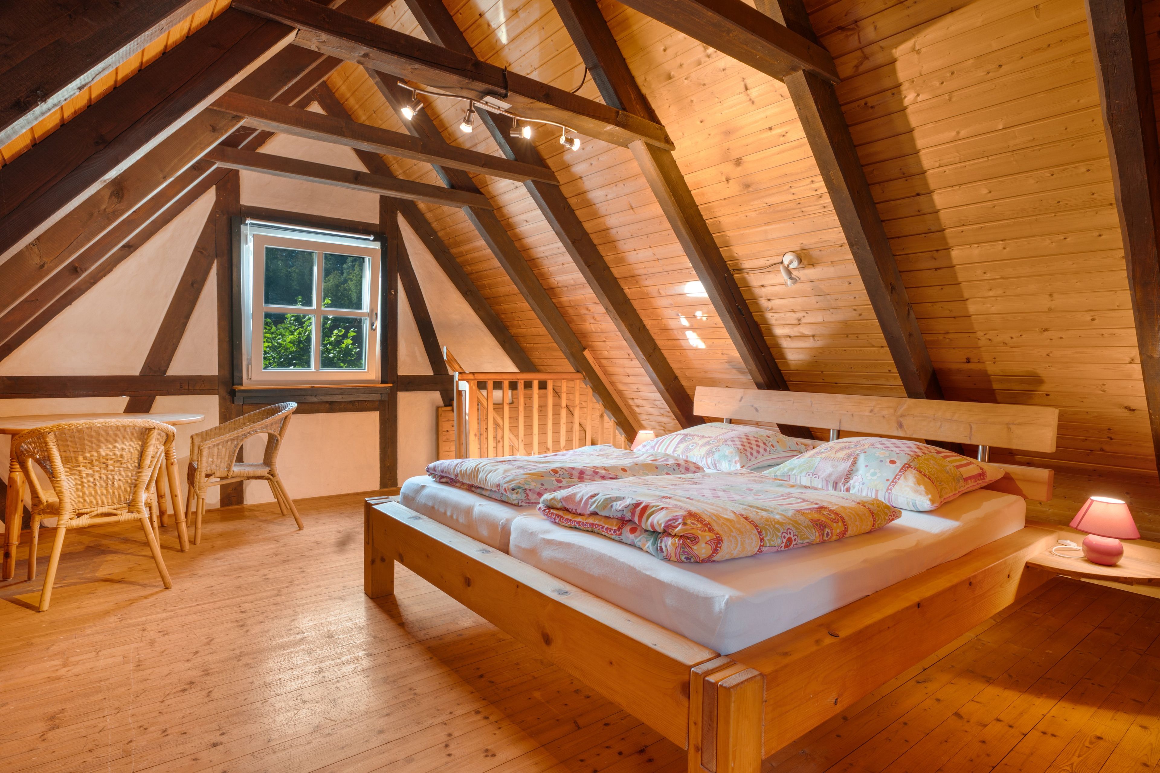 Un dormitorio acogedor en el ático, con techo de madera, cama con ropa de cama estampada, una mesita pequeña y una silla.