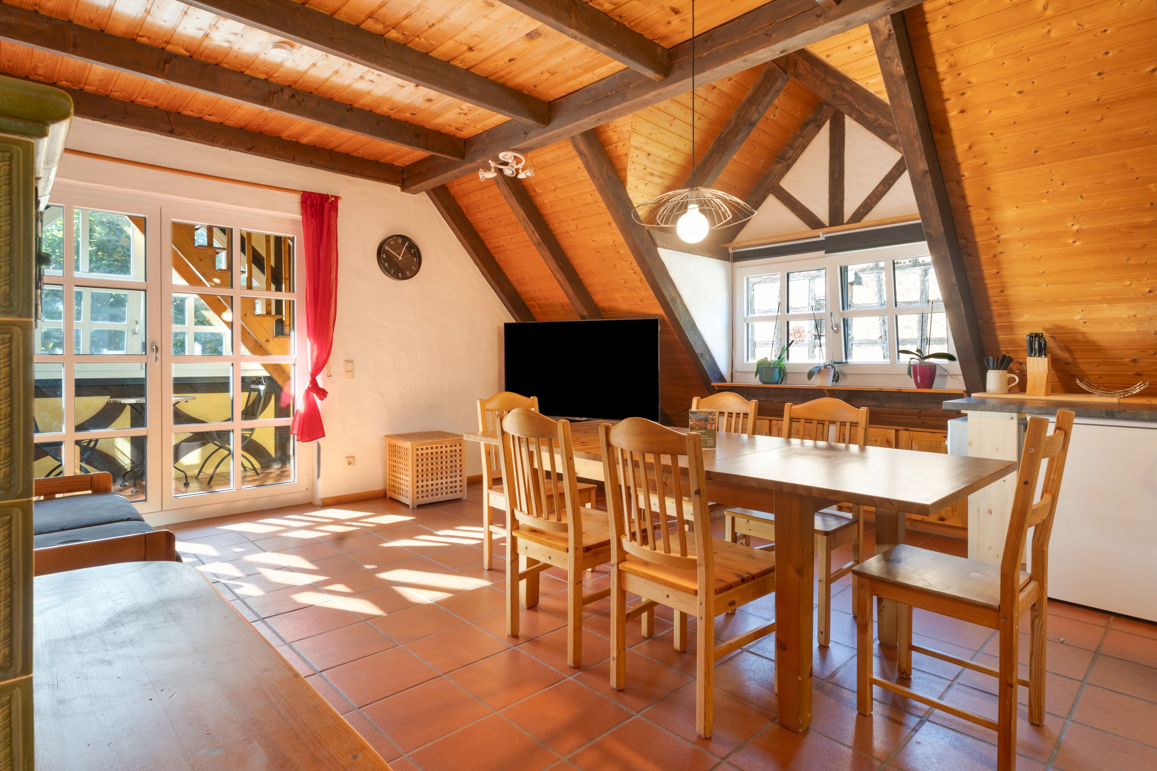 A dining area with a wooden table and chairs, a television, and a window with a red curtain.