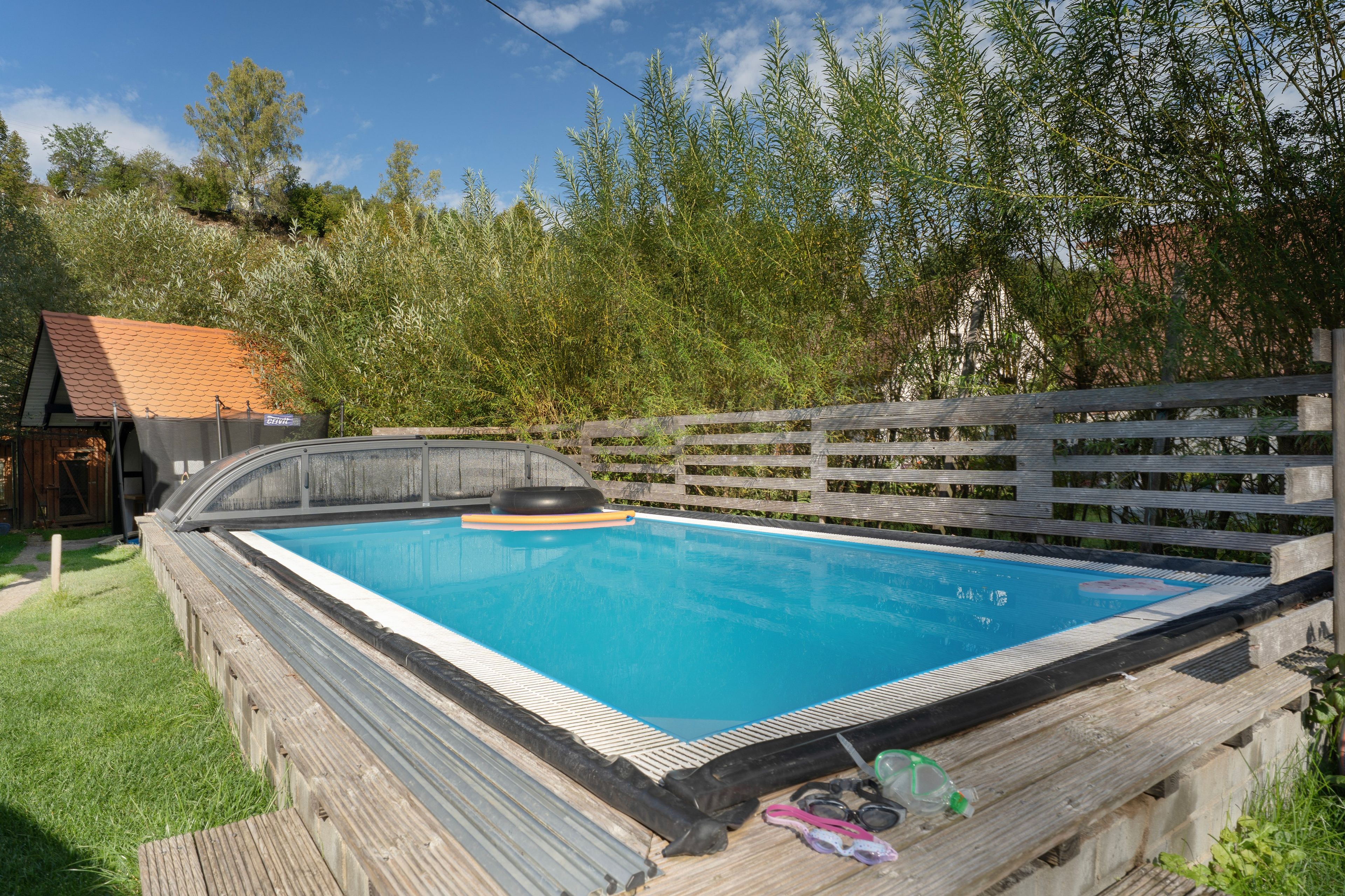 A rectangular swimming pool with a wooden deck surrounding it, a small shed in the background, and a wooden fence with a statue.