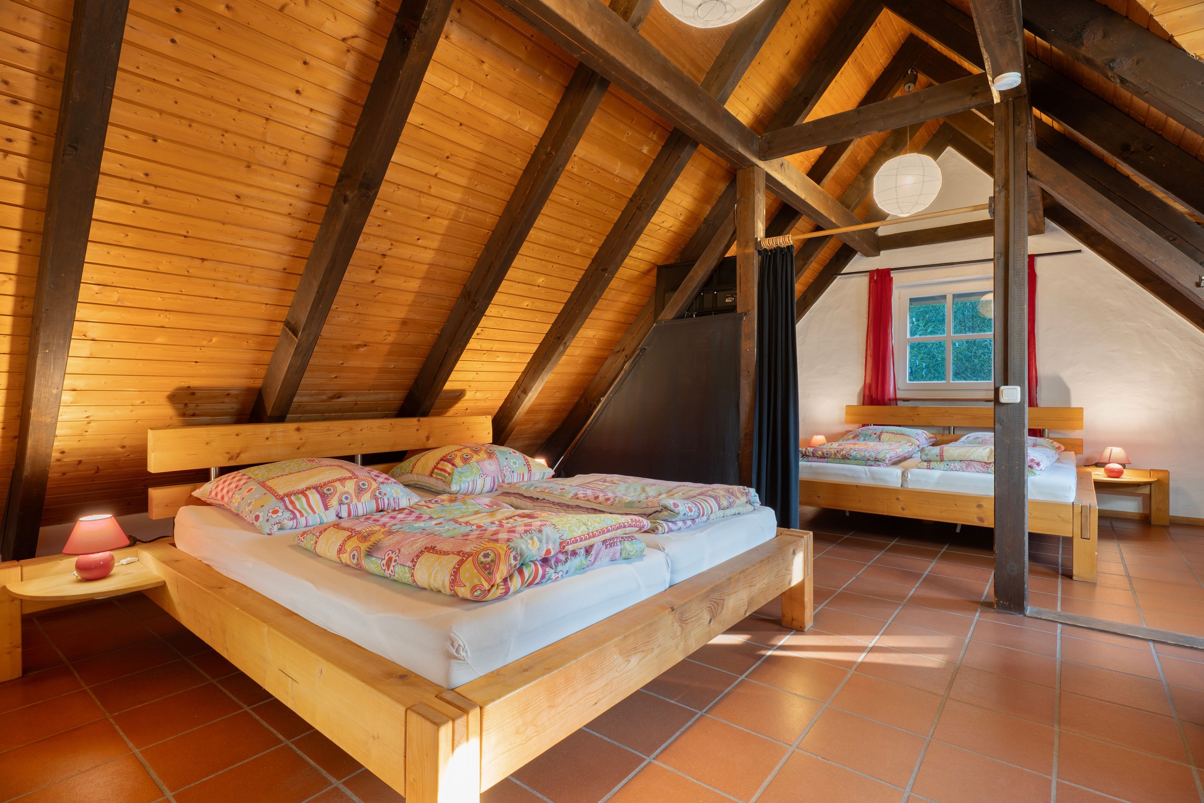Une chambre confortable située dans les combles, avec un plafond en bois, un grand lit recouvert d’une literie colorée, une table de chevet avec une lampe et une fenêtre agrémentée de rideaux.