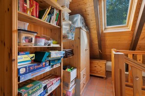 Un intérieur en bois avec un plafond en pente, des étagères en bois remplies de livres et de jeux, une fenêtre donnant sur des arbres et un escalier en bois.