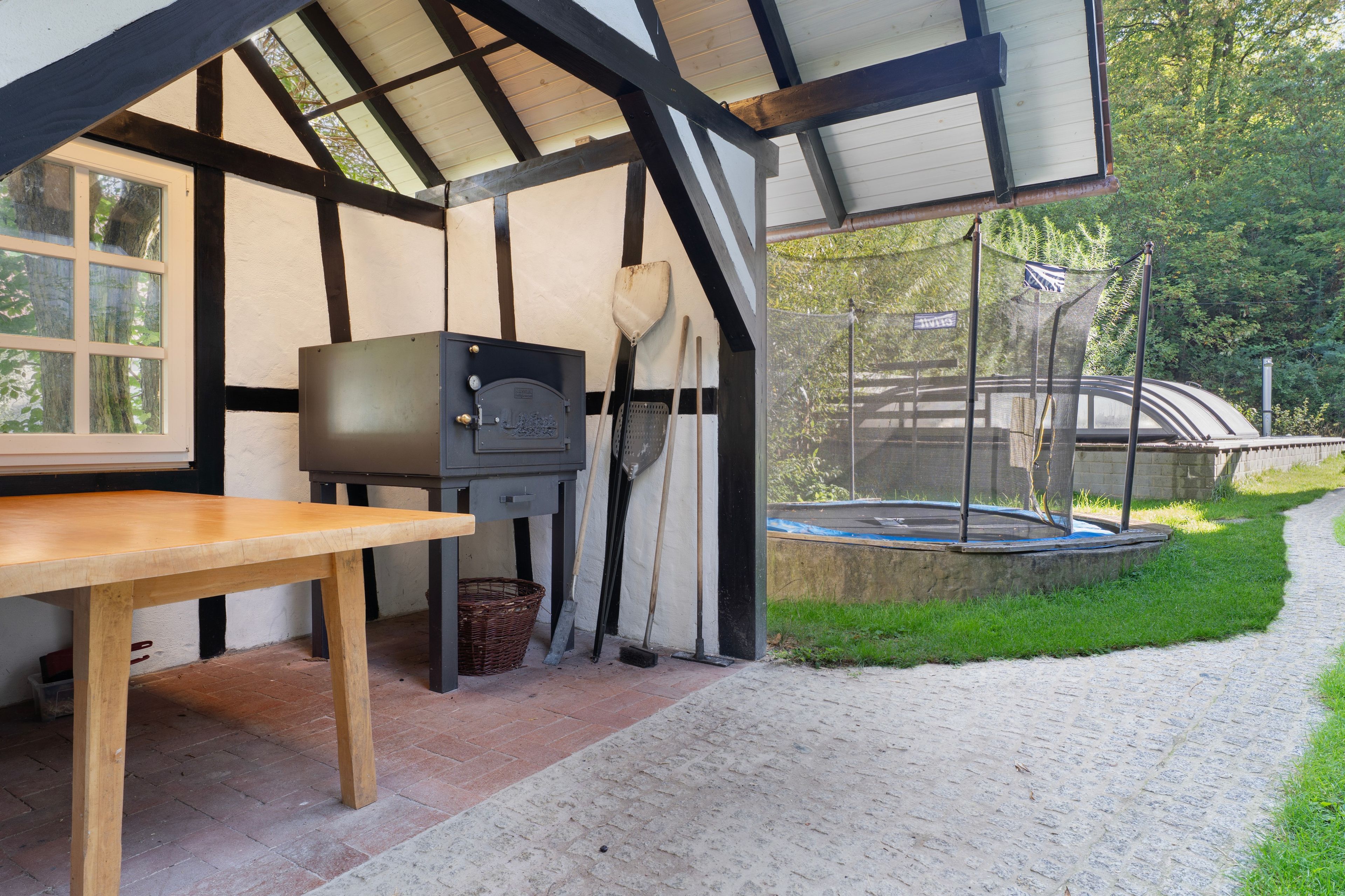 A covered outdoor area with a table and a grill, surrounded by a garden and a stone wall.