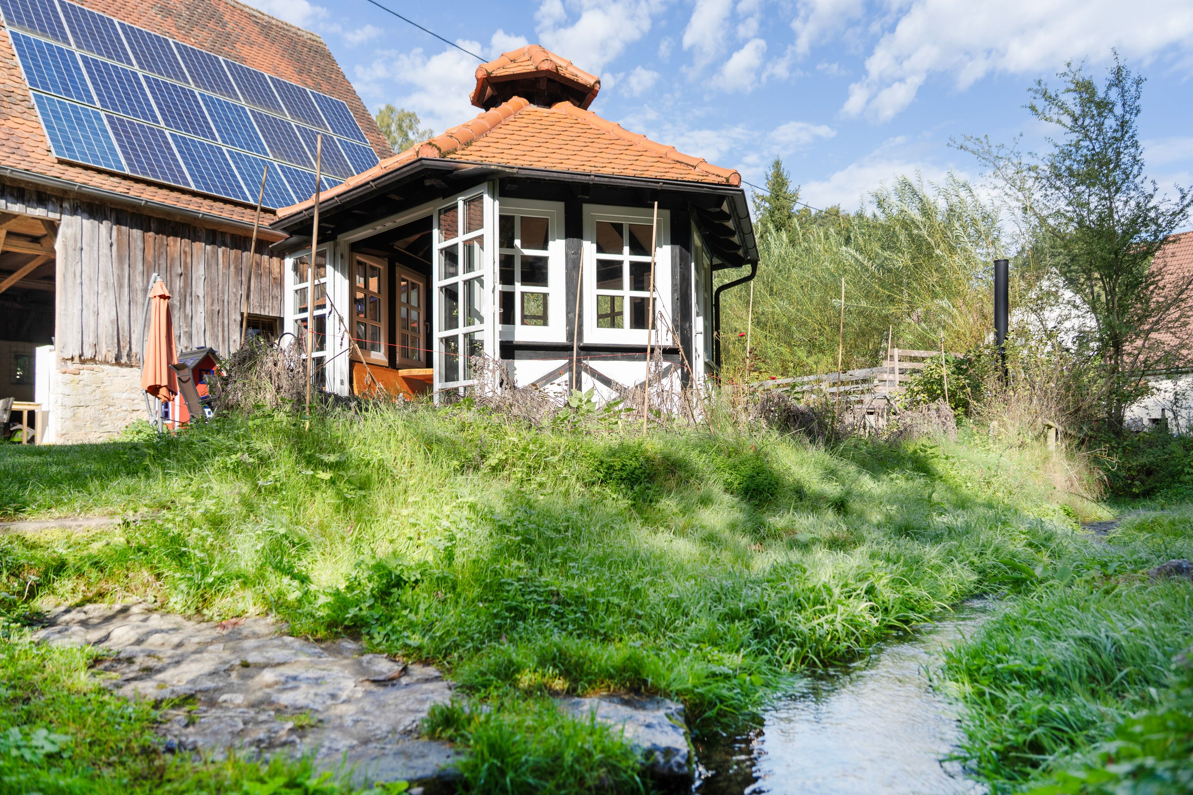 Een landelijke woning met een zonnepaneelinstallatie op het dak, een kleine tuin en een smalle beek.